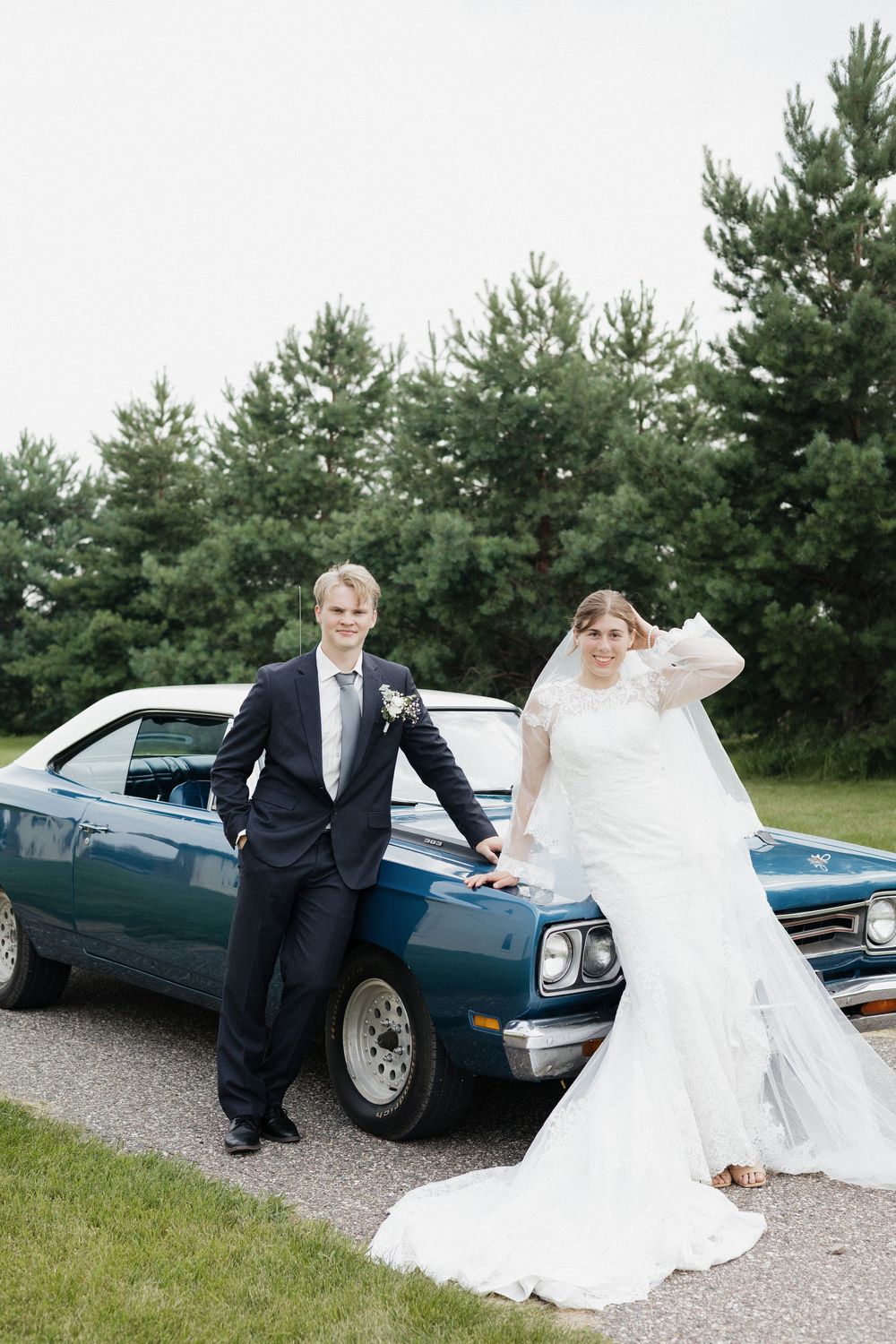 A classic car serves as a backdrop for wedding photos on a gravel road with pine trees in the background.