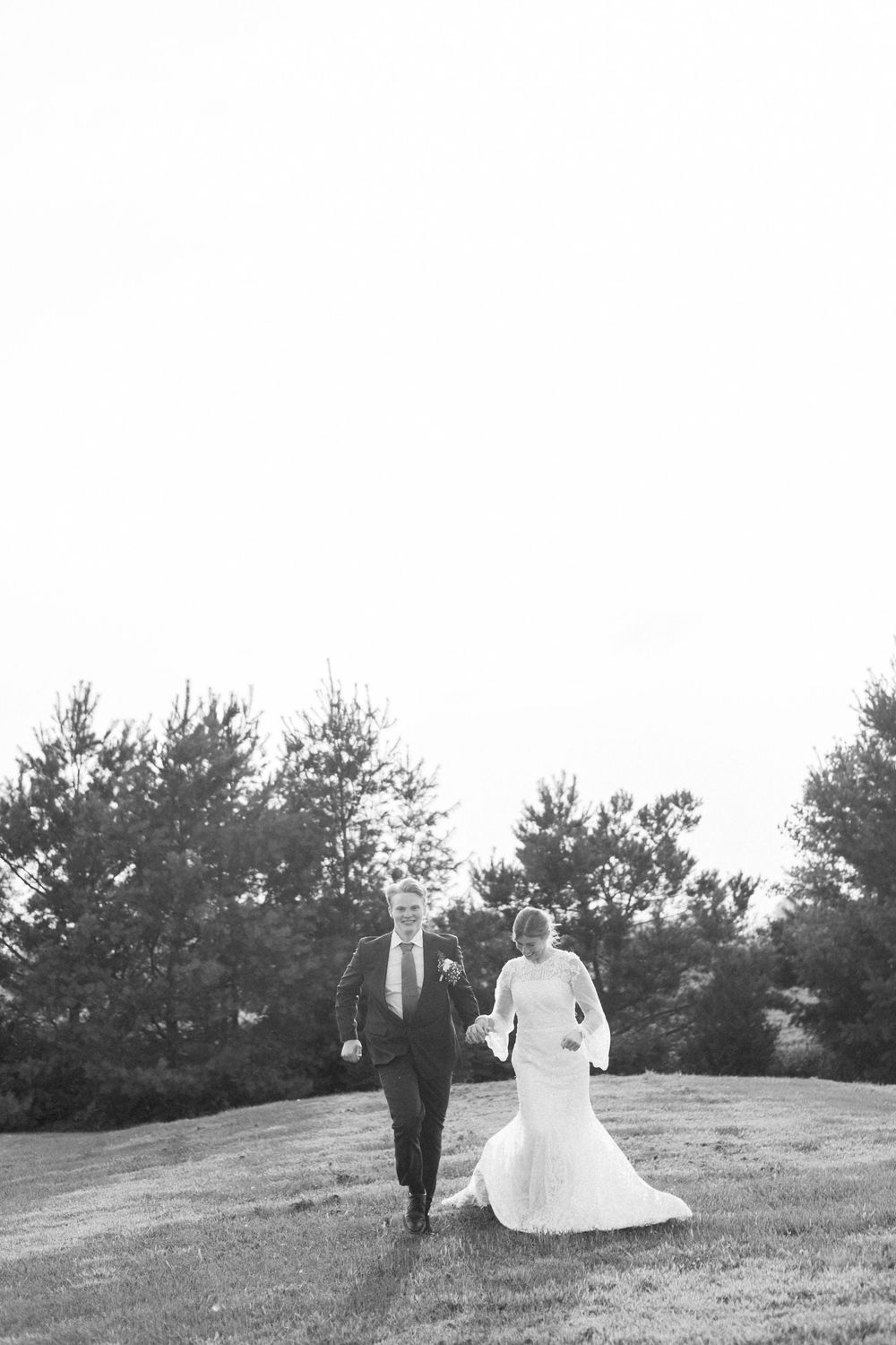 A couple walks through a grassy hillside on their wedding day with evergreen trees in the distance.