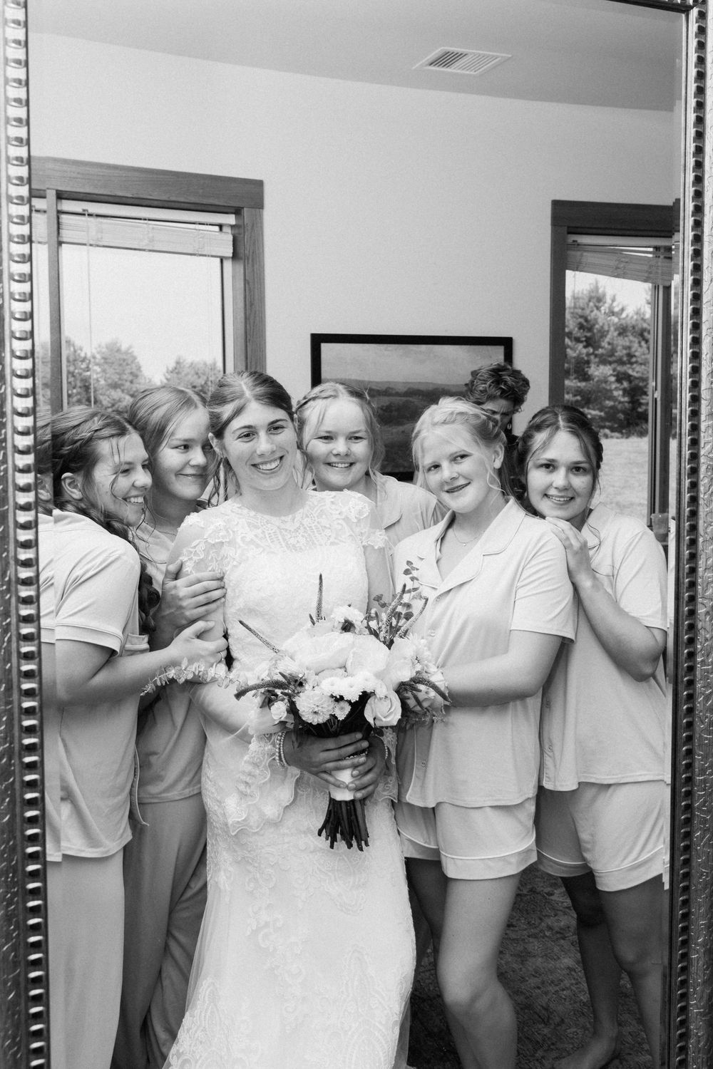 A joyful black and white photo of a bridal party gathering around a mirror with flowers.