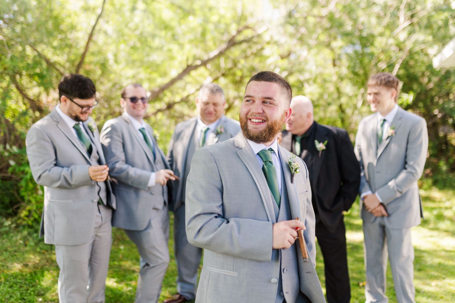 Wedding party in light gray suits and green boutonnieres gather outdoors beneath trees in sunlight.