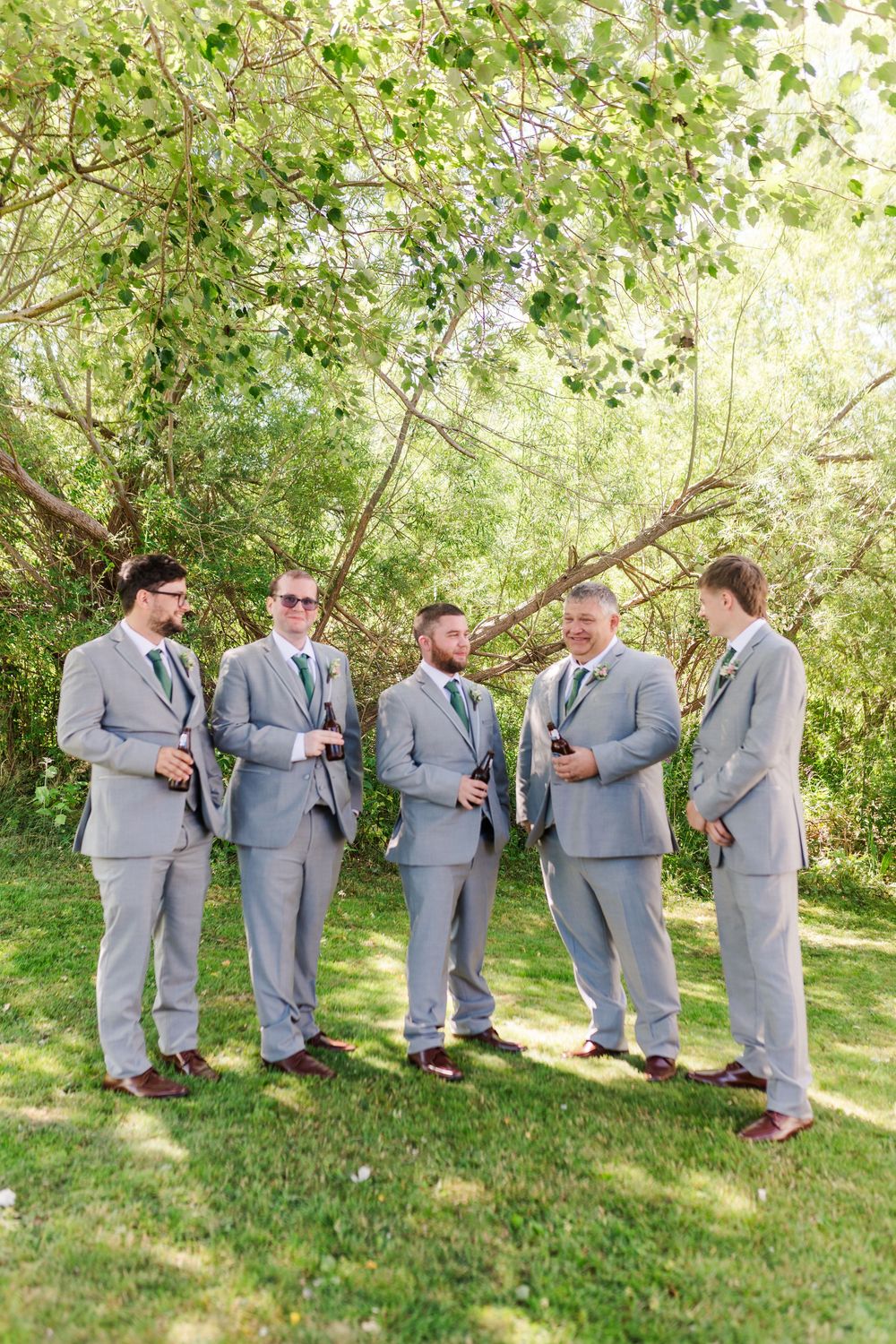 Groomsmen in matching light gray suits stand together on grass under trees having a casual conversation.