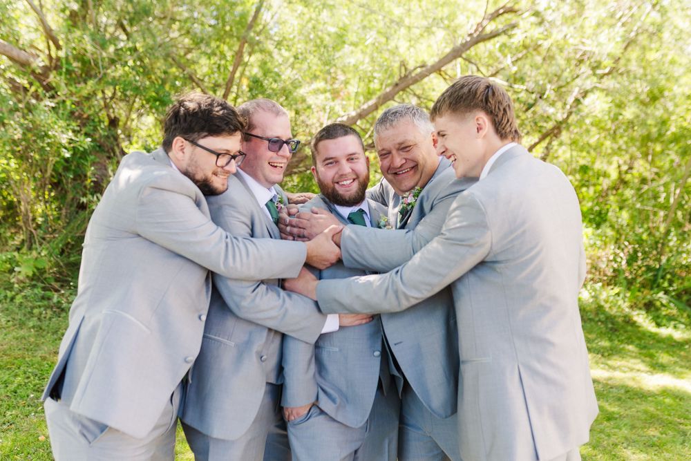 A group wearing light blue and gray suits embrace in a casual outdoor setting with green foliage in the background.