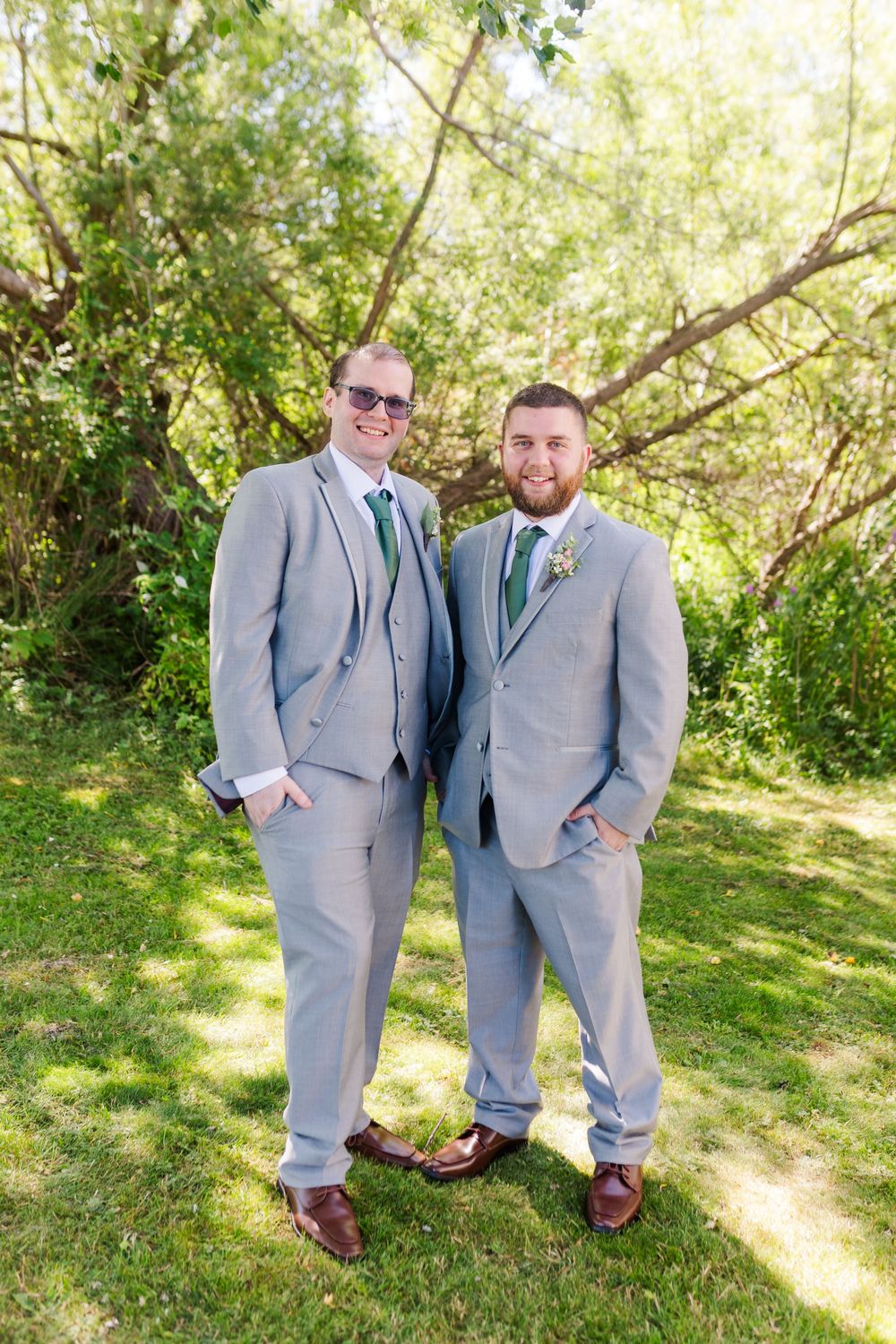 Groomsmen in grey suits posing together on grassy hill with trees in background.
