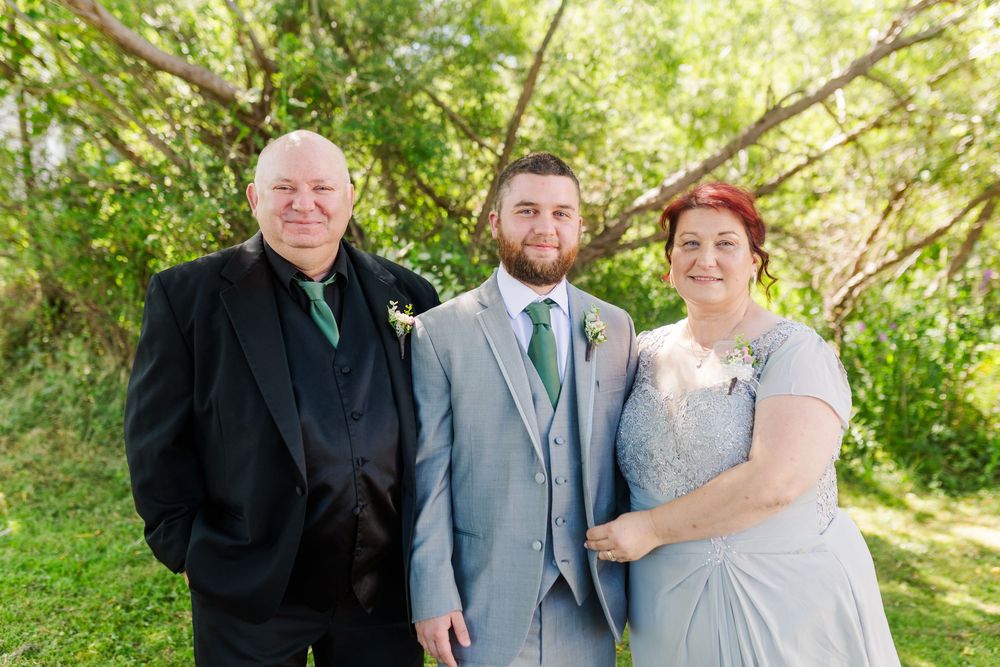 Wedding party members wearing gray suits and formal attire pose together in a sunny outdoor garden setting.