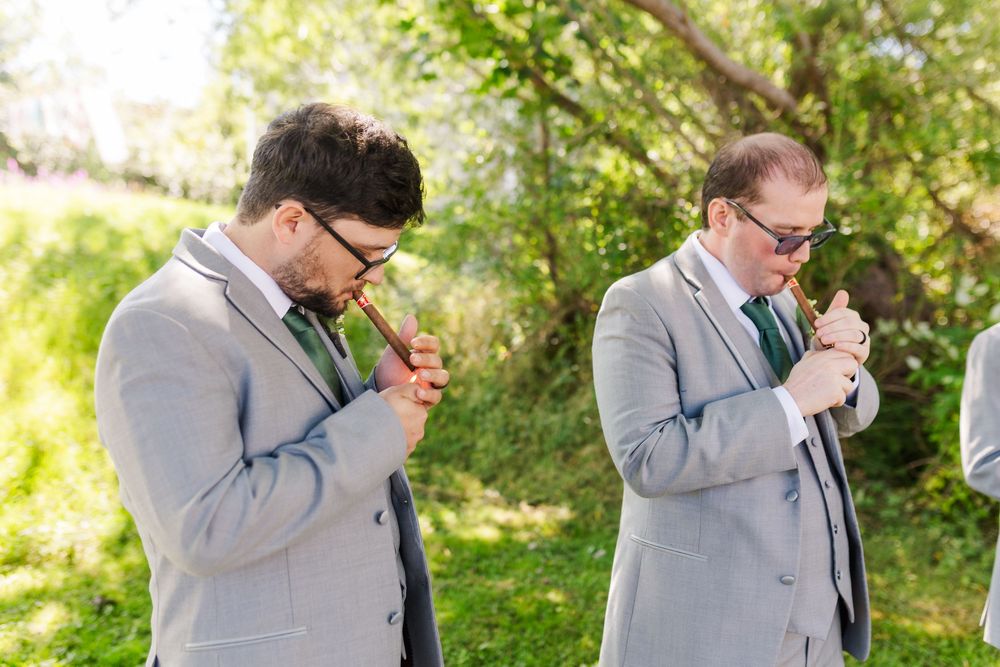 Two men in gray suits lighting cigars together in a sunny outdoor garden setting.