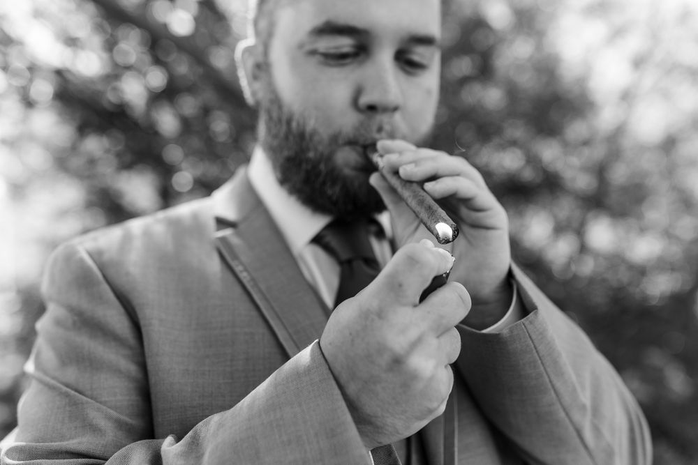 Groomsmen in light gray suits share cigars and laugh together outdoors during a wedding celebration.