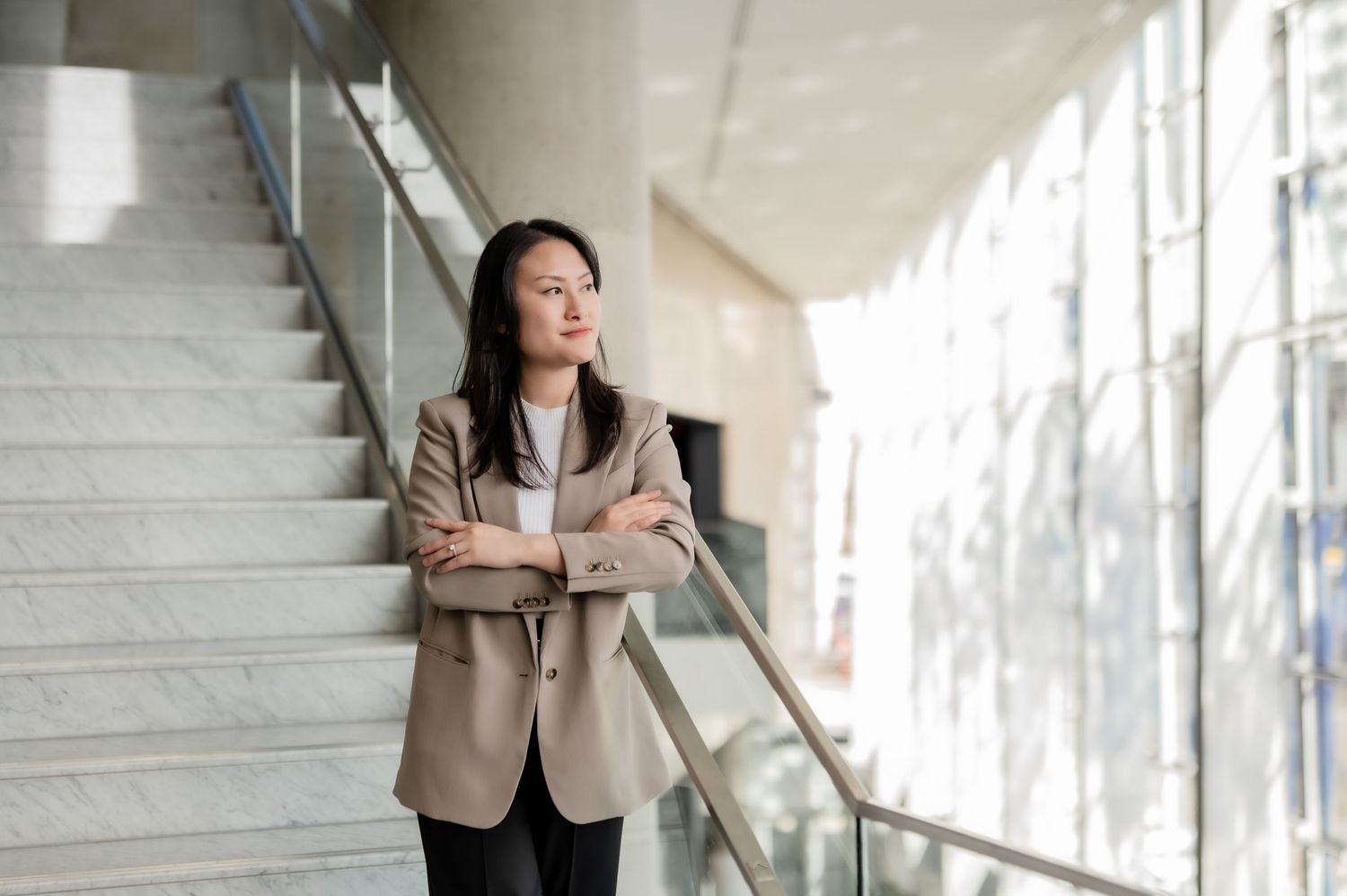 Professional dressed figure standing in modern bright lobby with glass windows and staircase behind.