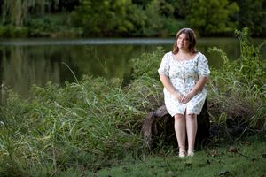 Woman in white dress sitting on grass near a pond in a tranquil outdoor setting.