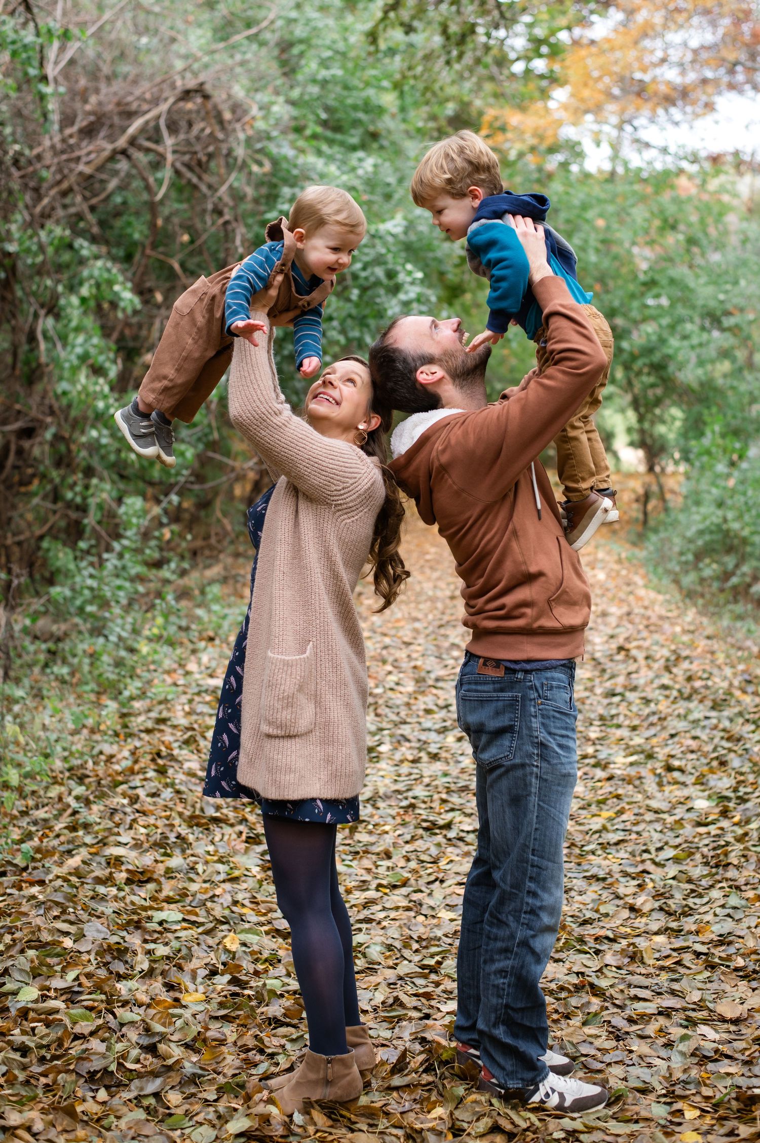 A couple playfully lifts young children in the air on a leaf-covered autumn forest path.