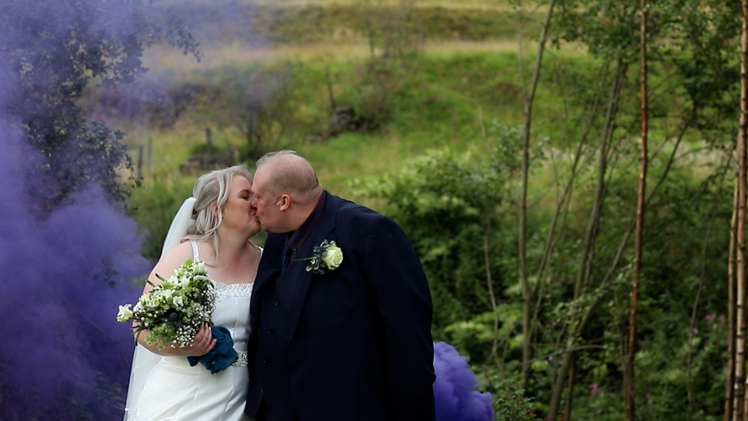 A wedding couple embraces against a backdrop of purple smoke and green countryside.