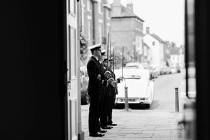 A series of silhouetted figures stand against an old city street with white stone buildings in black and white photography.
