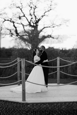 Three black and white romantic photos of a couple in formal wedding attire standing on a wooden dock at dusk.