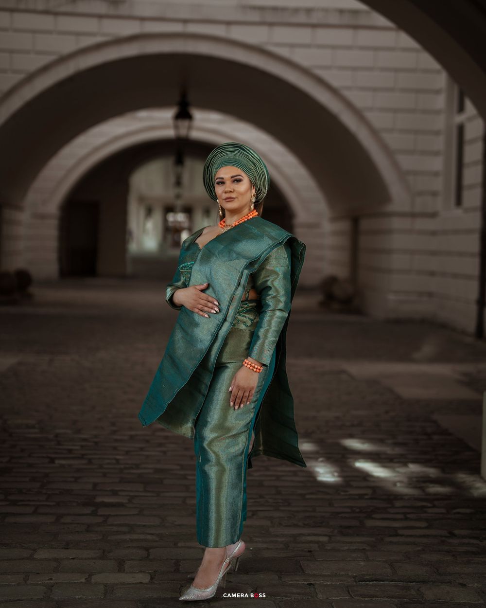 Woman in elegant emerald green dress poses dramatically in an arched corridor with cobblestone flooring.