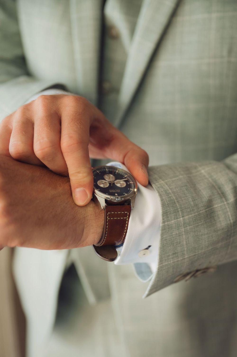 Close-up of hands adjusting a brown leather watch strap on gray suit sleeve.