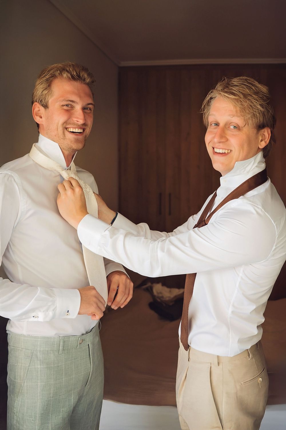 Two people playfully helping each other adjust their white dress shirts before a formal event.