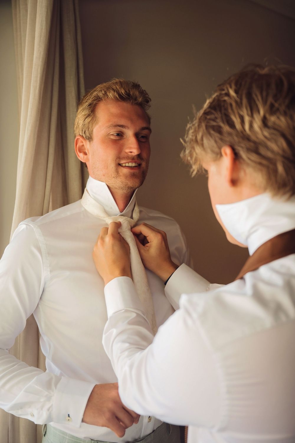 Friends adjusting each other's collars and ties while getting ready in formal white attire.