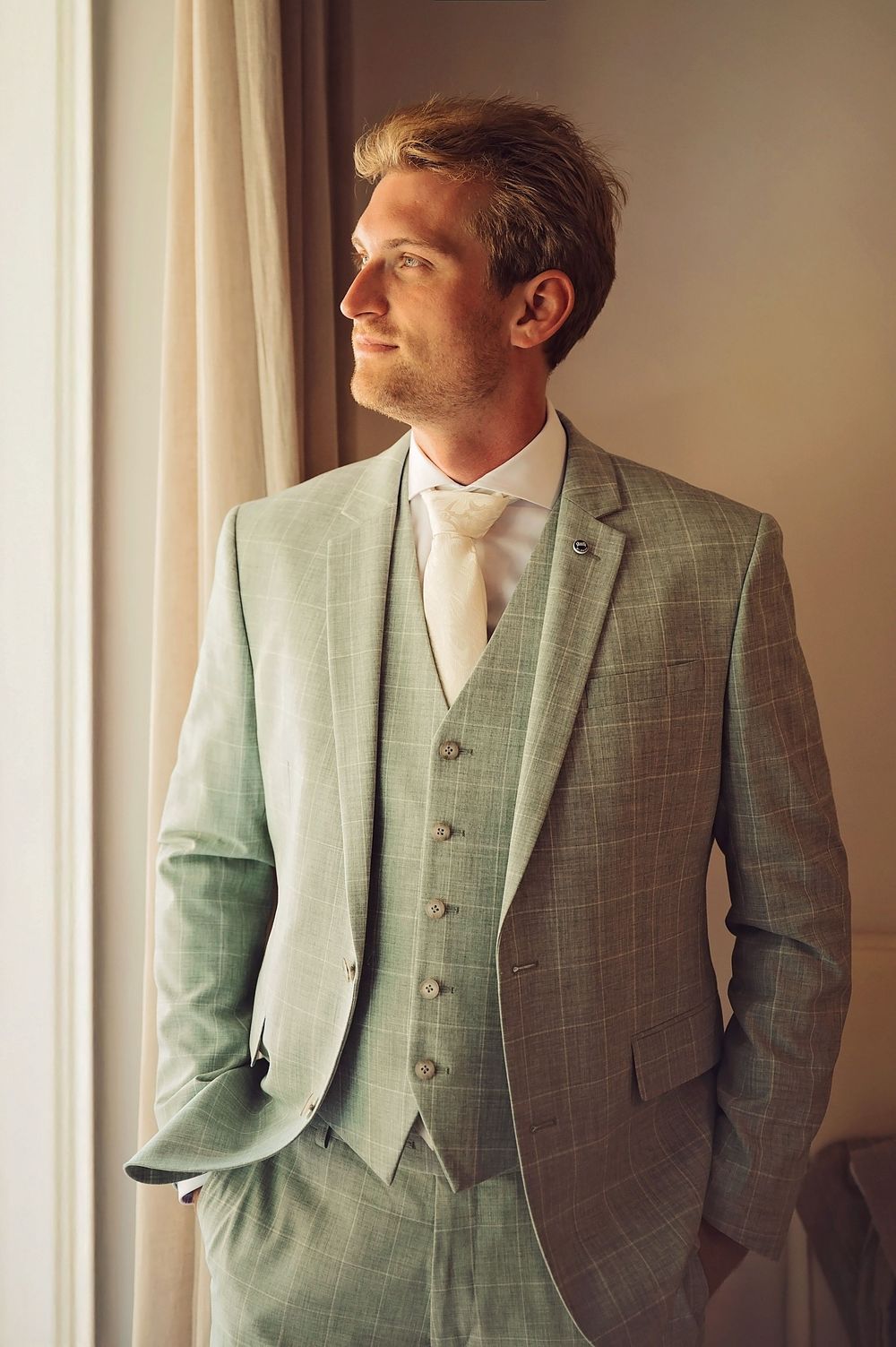 A groom in a light grey three-piece suit standing by a window in natural light.