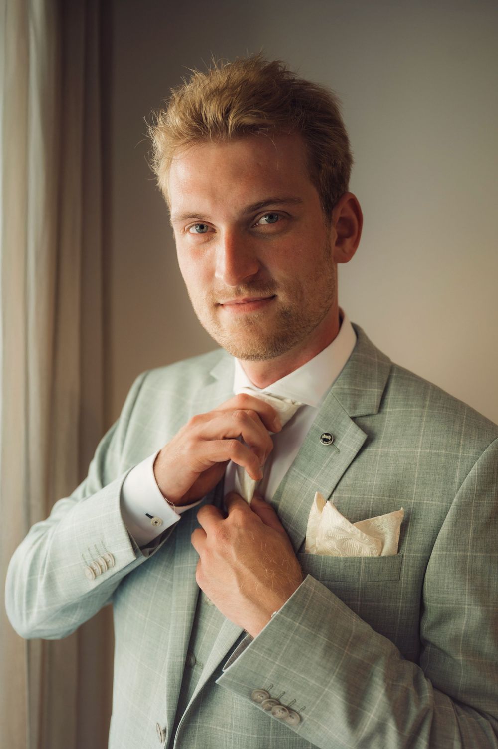 A groom adjusting his tie while wearing a light grey checkered suit near a window.