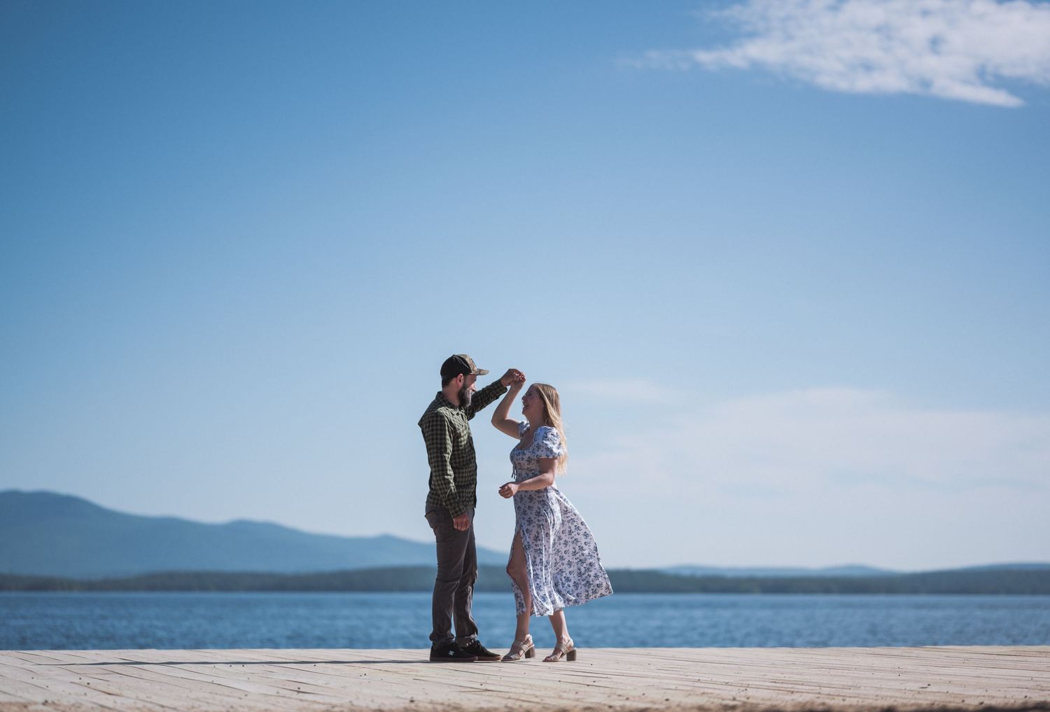 Couple dancing on a wooden dock by a lake under a bright blue sky with mountains in the distance.