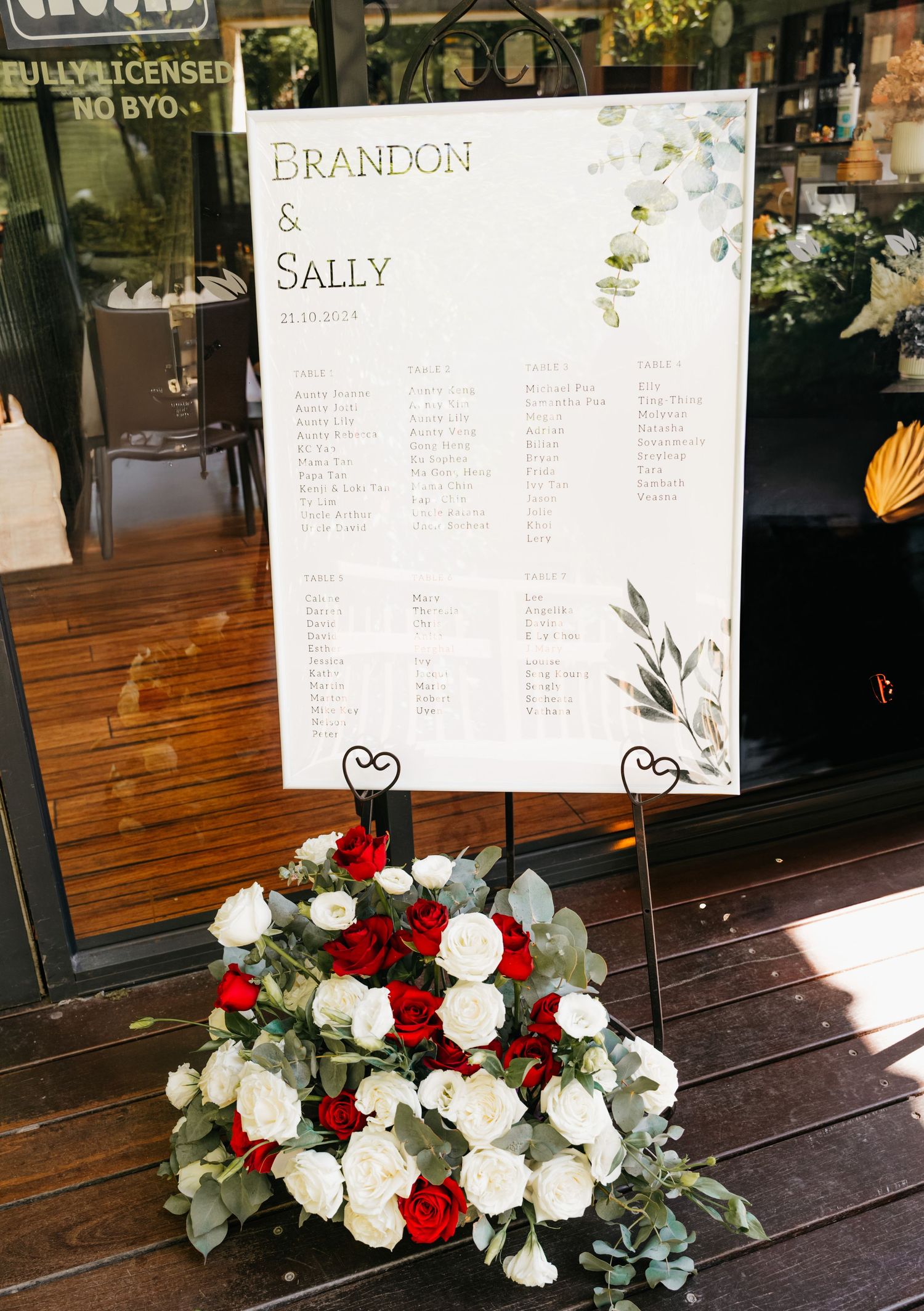 A wedding seating chart display with floral decorations featuring white and red roses on a wooden table.