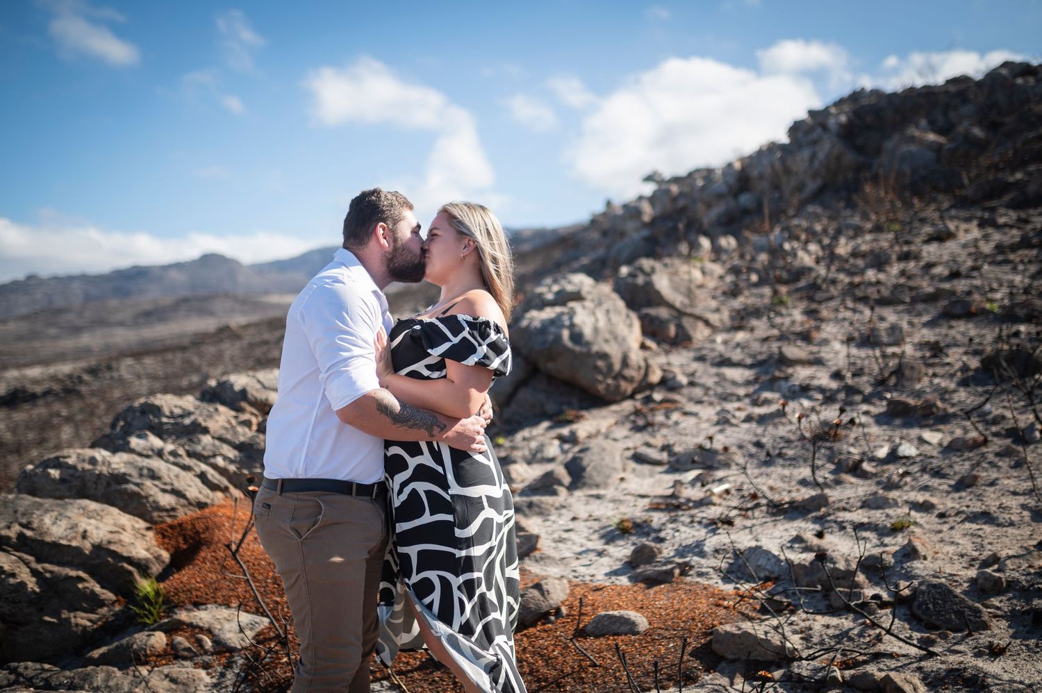 A couple embraces while sitting on rugged desert rocks against a scenic mountain backdrop.