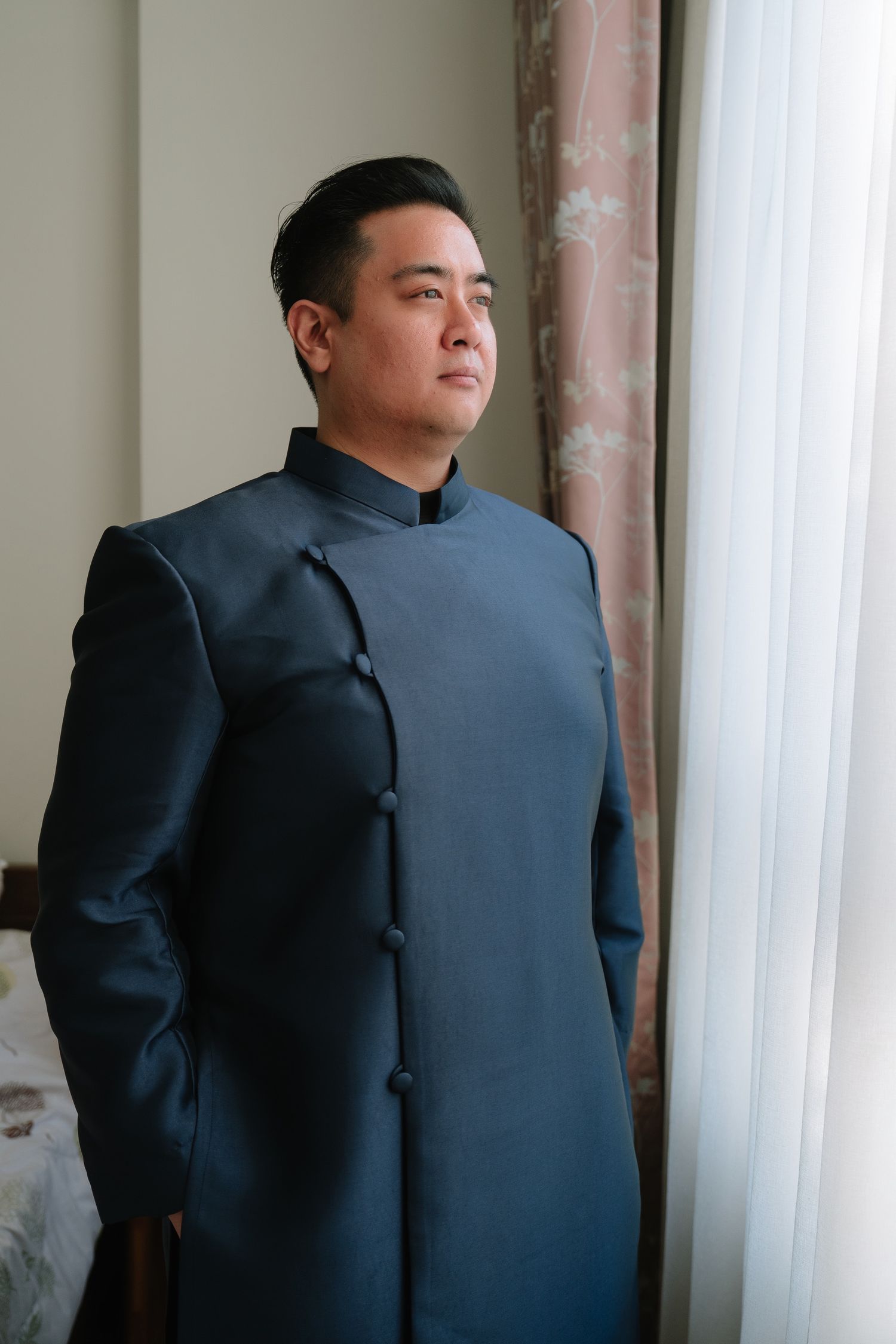 Asian groom in a dark blue mandarin collar wedding suit standing by a window.
