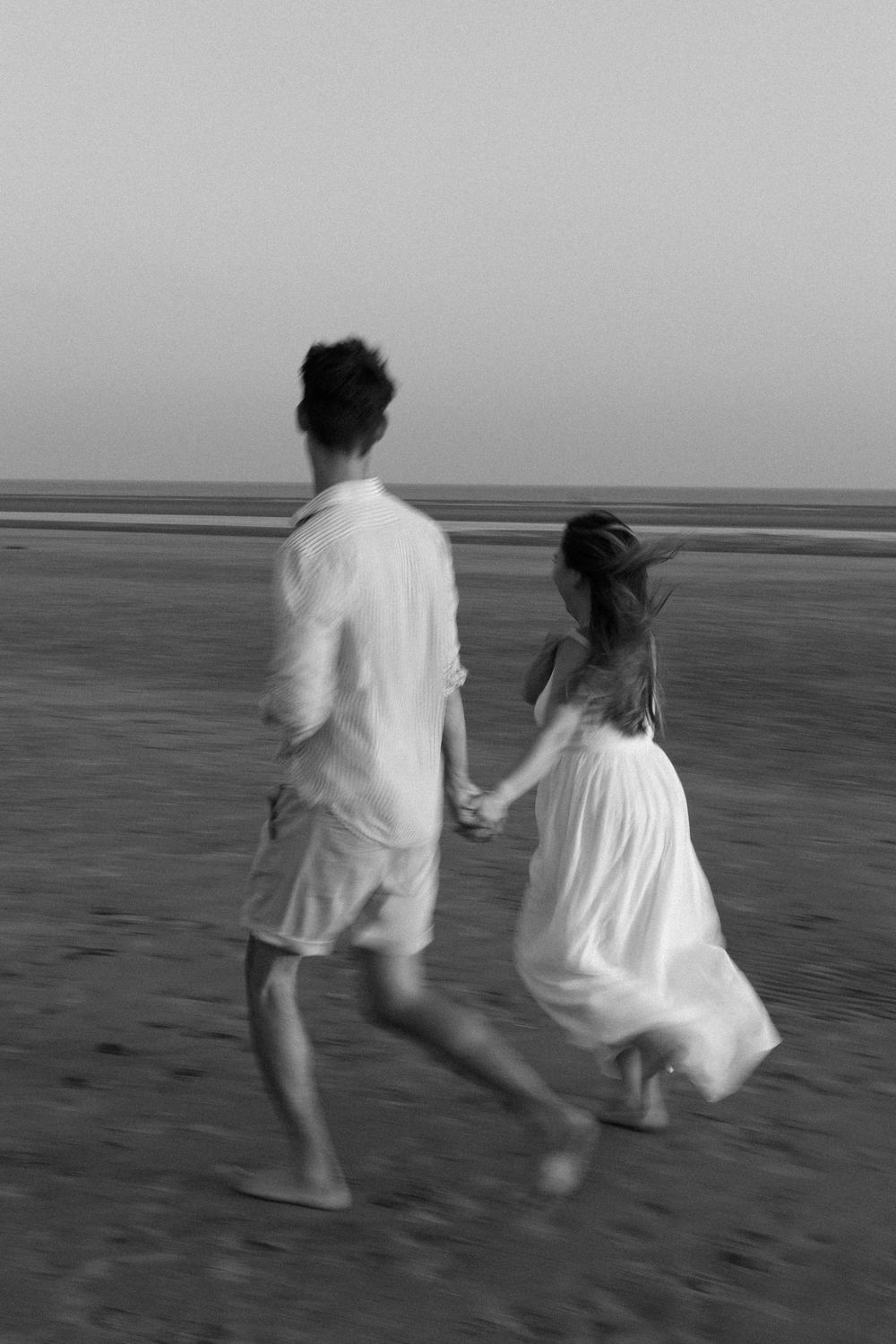 Couple in white clothing running joyfully along the beach at dusk in black and white.