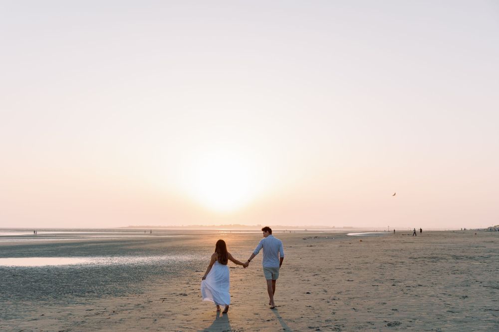 Two silhouettes walk hand in hand along a beach during sunset.
