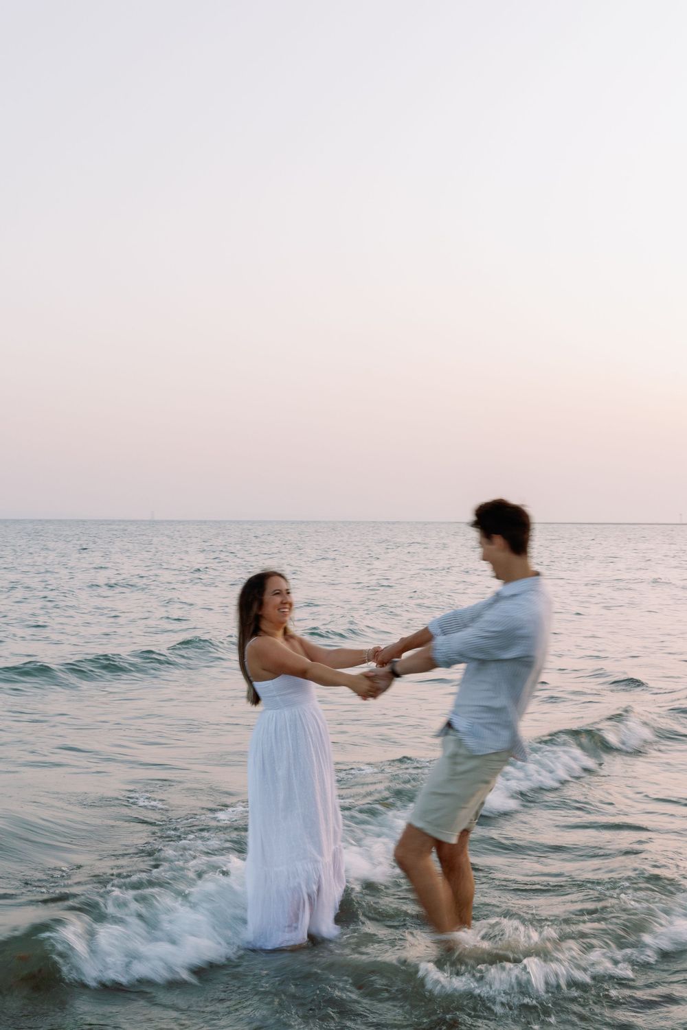 Playful couple splashes and dances in ocean waves during sunset wearing white and blue outfits.