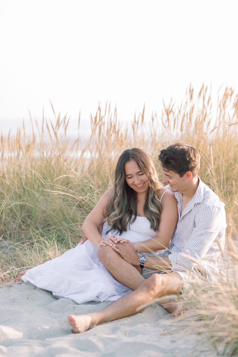 Couple relaxing together on sandy beach with tall golden grass in background during sunset.