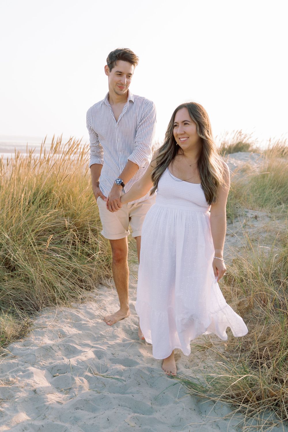 A romantic couple in white outfits walking barefoot through sandy beach dunes.
