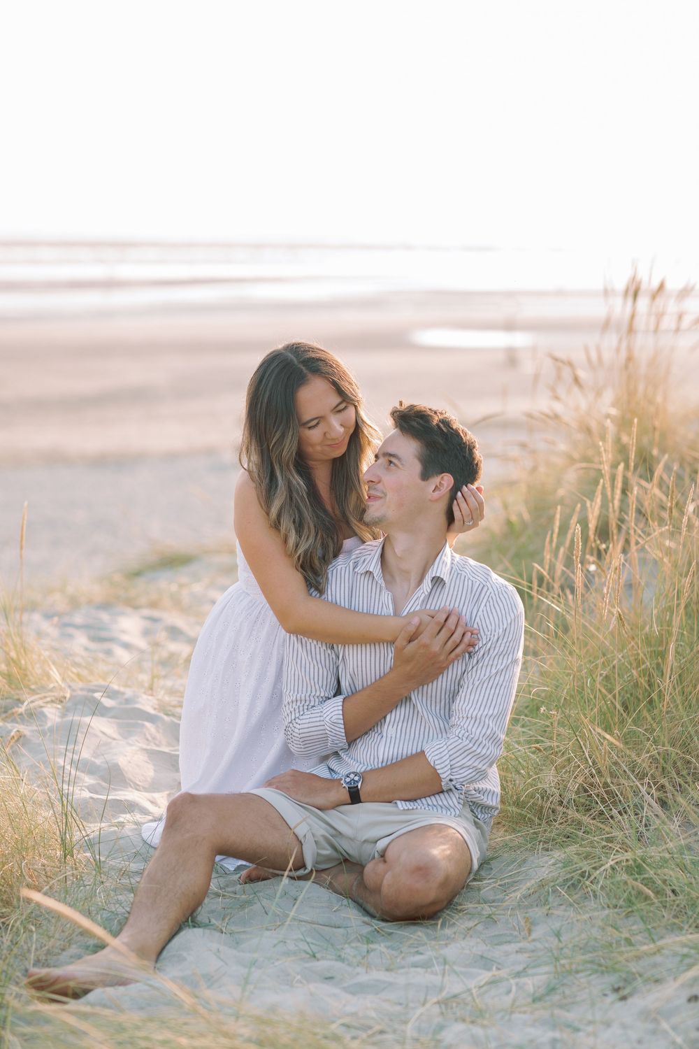 Romantic couple lounging on sandy beach dunes with tall grass at golden hour.
