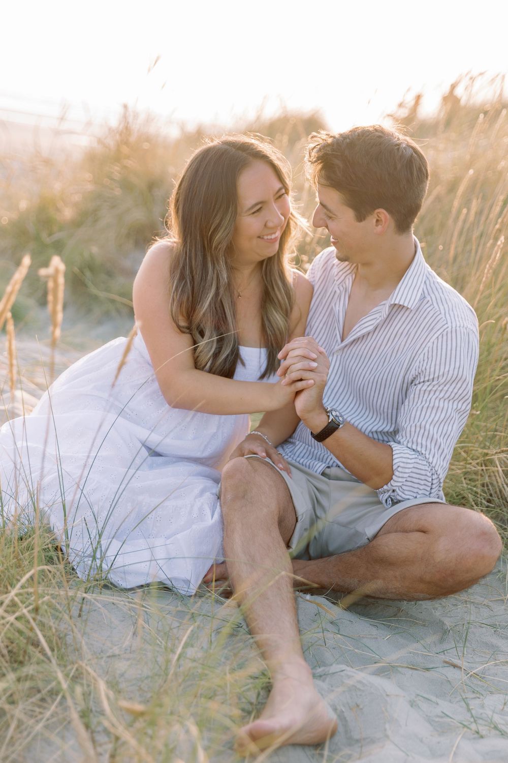 A couple shares an intimate moment together on a sandy beach at sunset among tall beach grass.