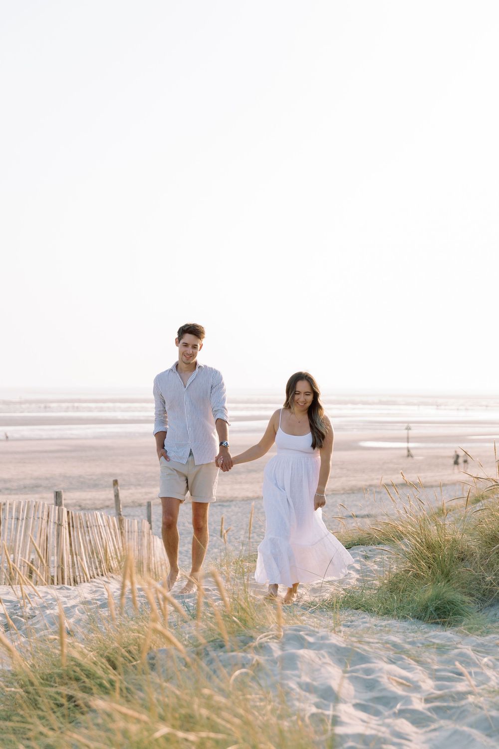 A couple in light summer attire walks together on a sandy beach at sunset among coastal grass.