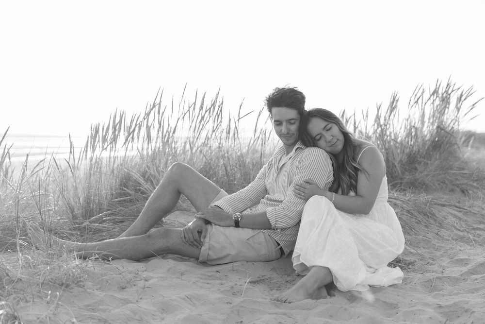 Couple lounging on sandy beach dunes with beach grass during sunset photo session.