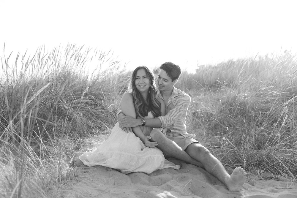 A couple lying in tall beach grass during golden hour at sunset.