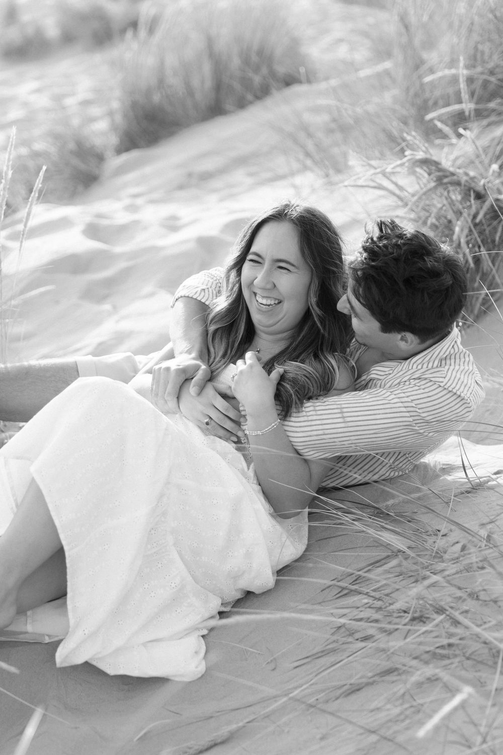 Black and white photo of couple lying together in beach grass sharing playful moments.