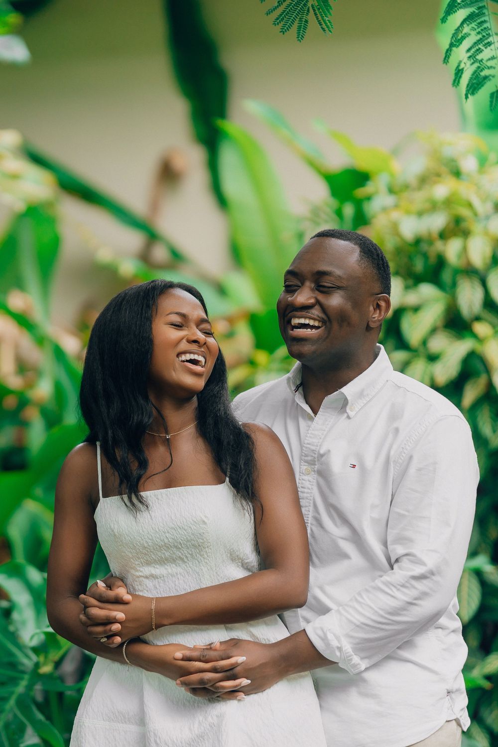 Happy couple in white outfits laughs and embraces while surrounded by lush green tropical plants.