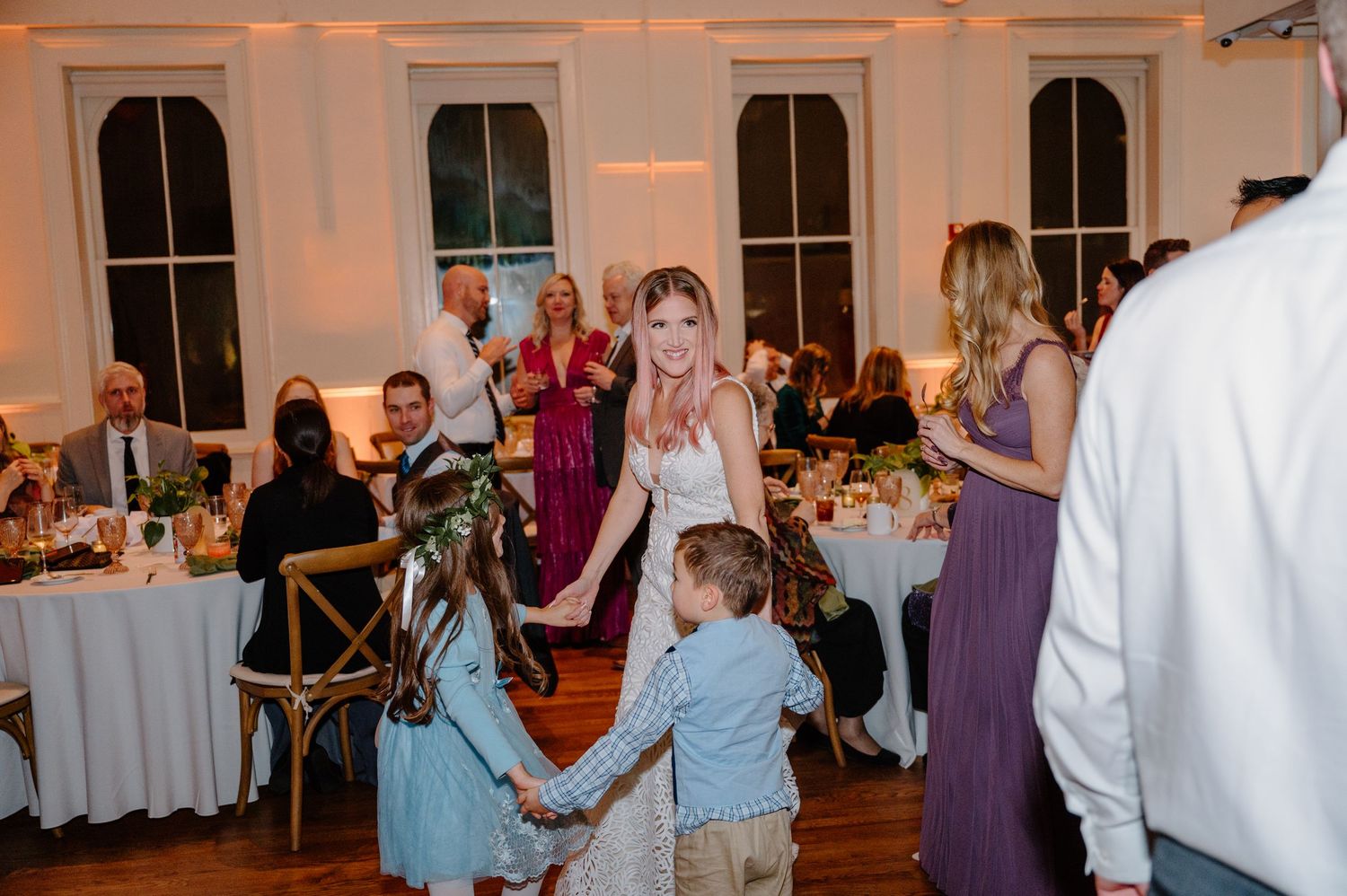 Wedding guests dance together on a wooden dance floor under string lights at an indoor reception venue.
