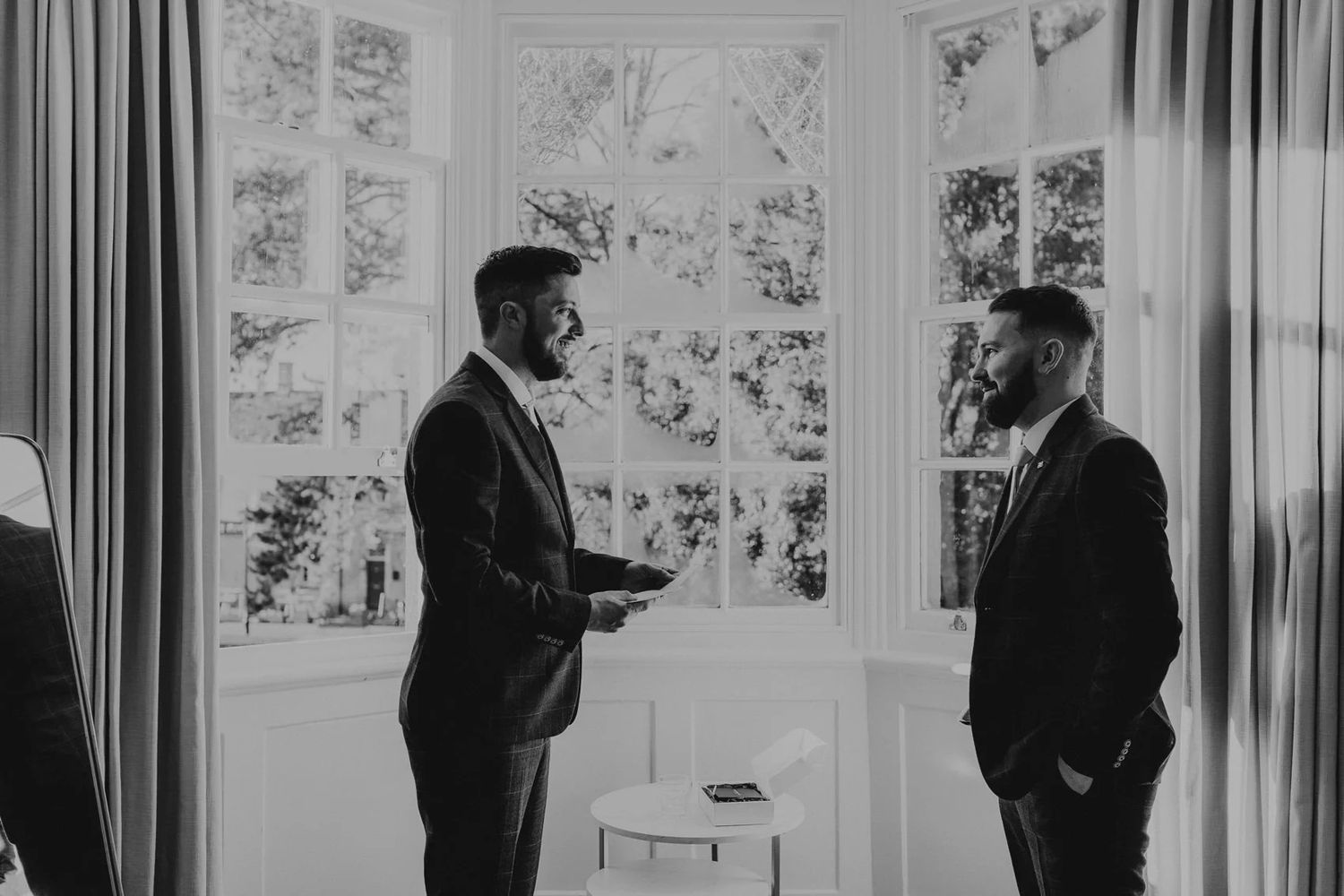 Black and white photo showing figures in conversation by large windows with curtains.