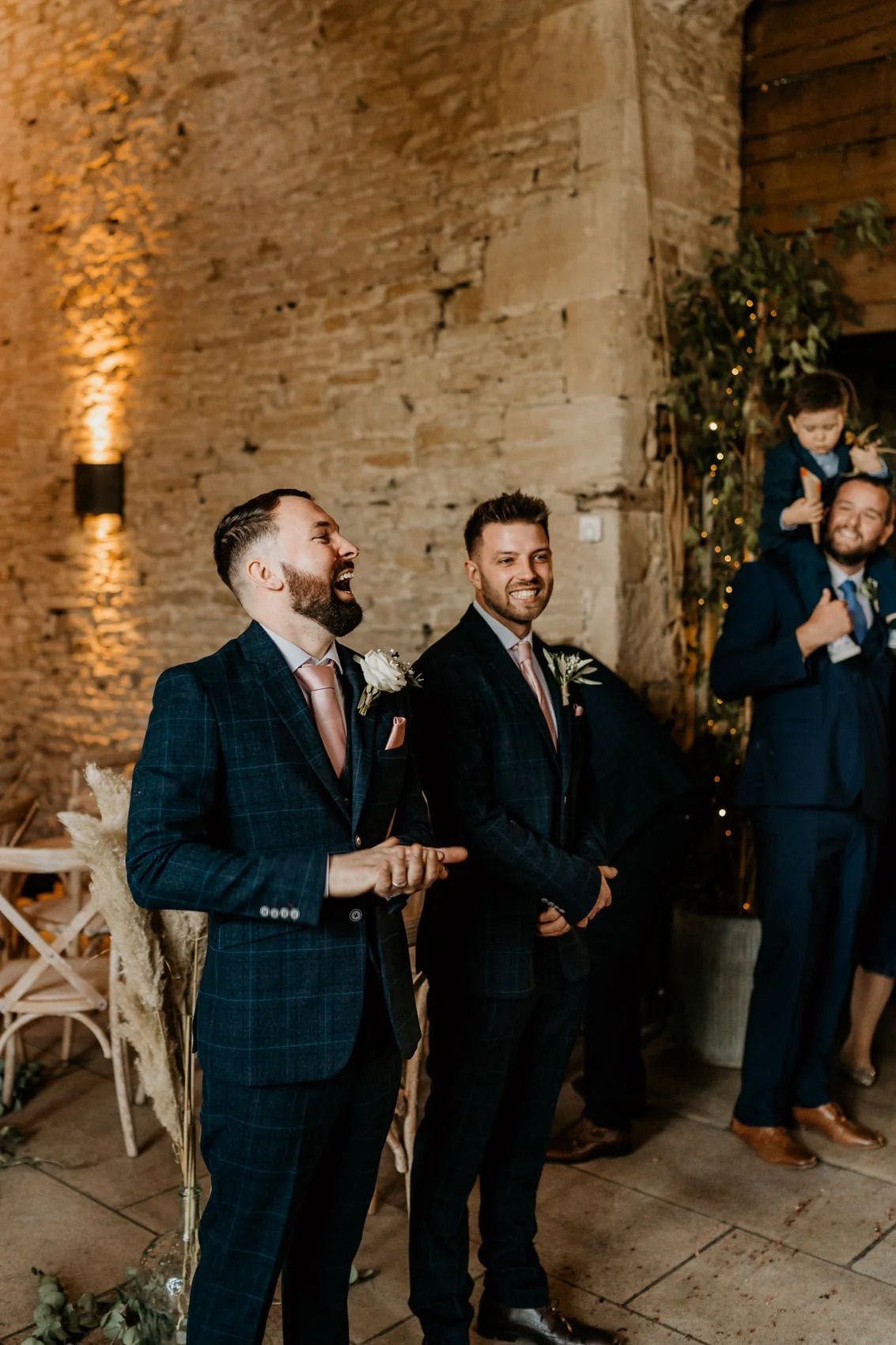 Groomsmen laughing together in dark suits at rustic brick venue.