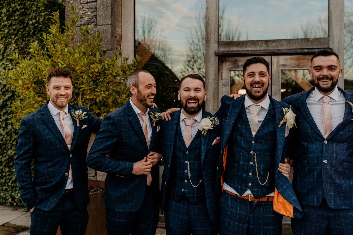 A group in navy suits stand together at a garden wedding celebration in autumn light.