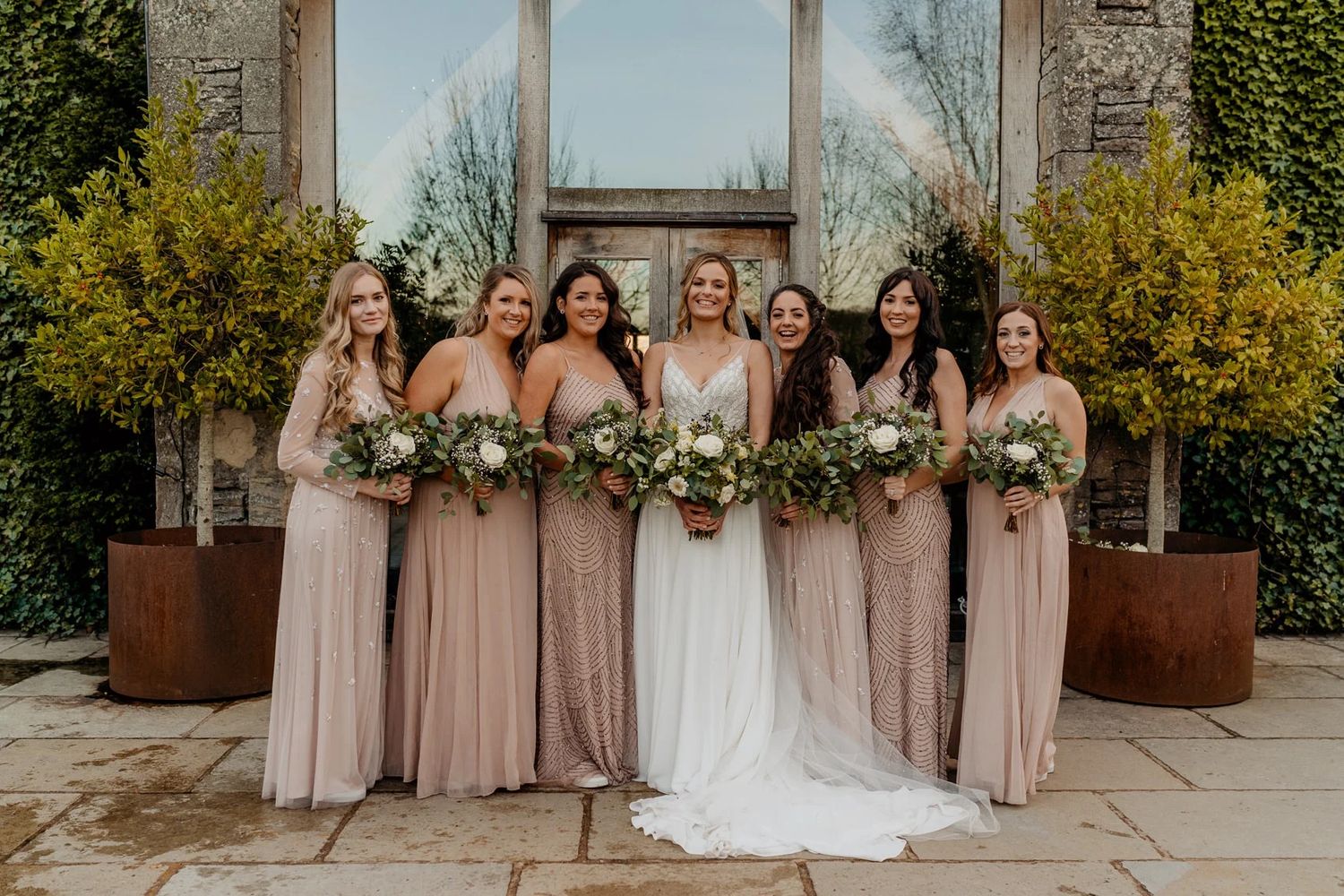 Bridal party stands together in elegant blush pink dresses against an outdoor stone and foliage backdrop.
