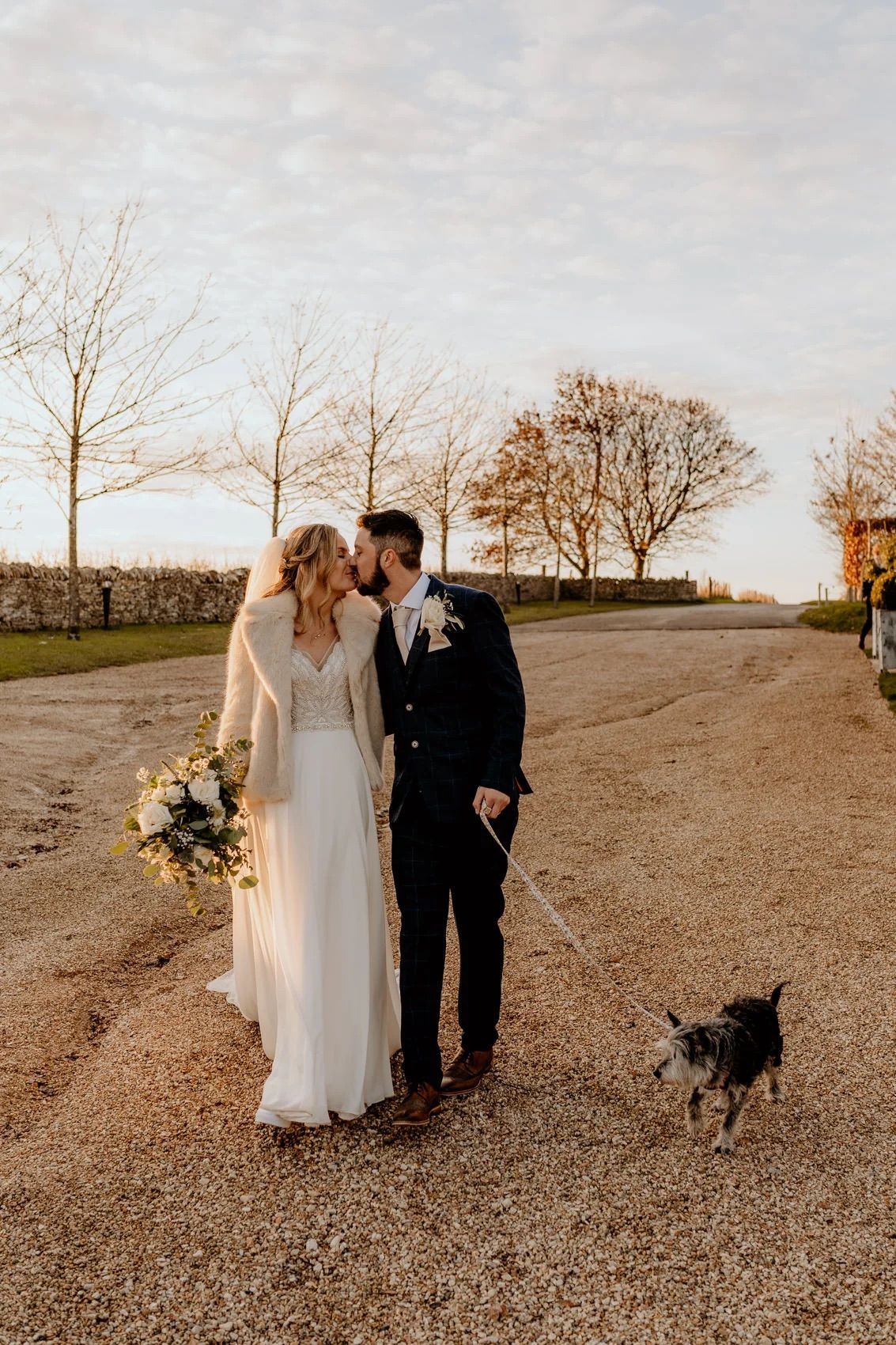 A couple shares an intimate moment in their wedding attire on a gravel path at sunset while their dog walks nearby.