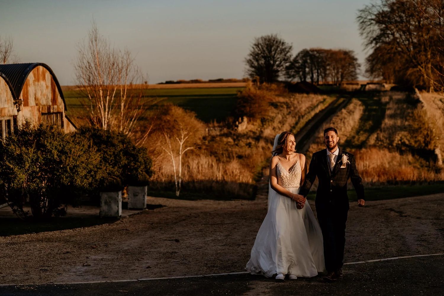 A couple in wedding attire strolls together during golden hour in a scenic hillside setting with autumn trees.