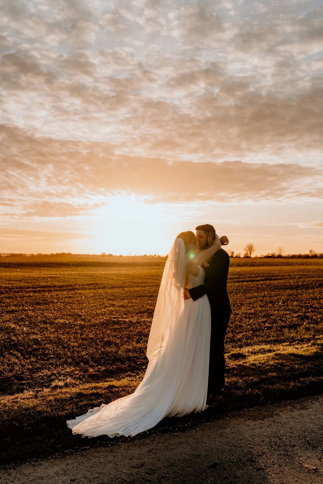 A couple in wedding attire embracing in a field at sunset with long shadows stretching across the golden landscape.