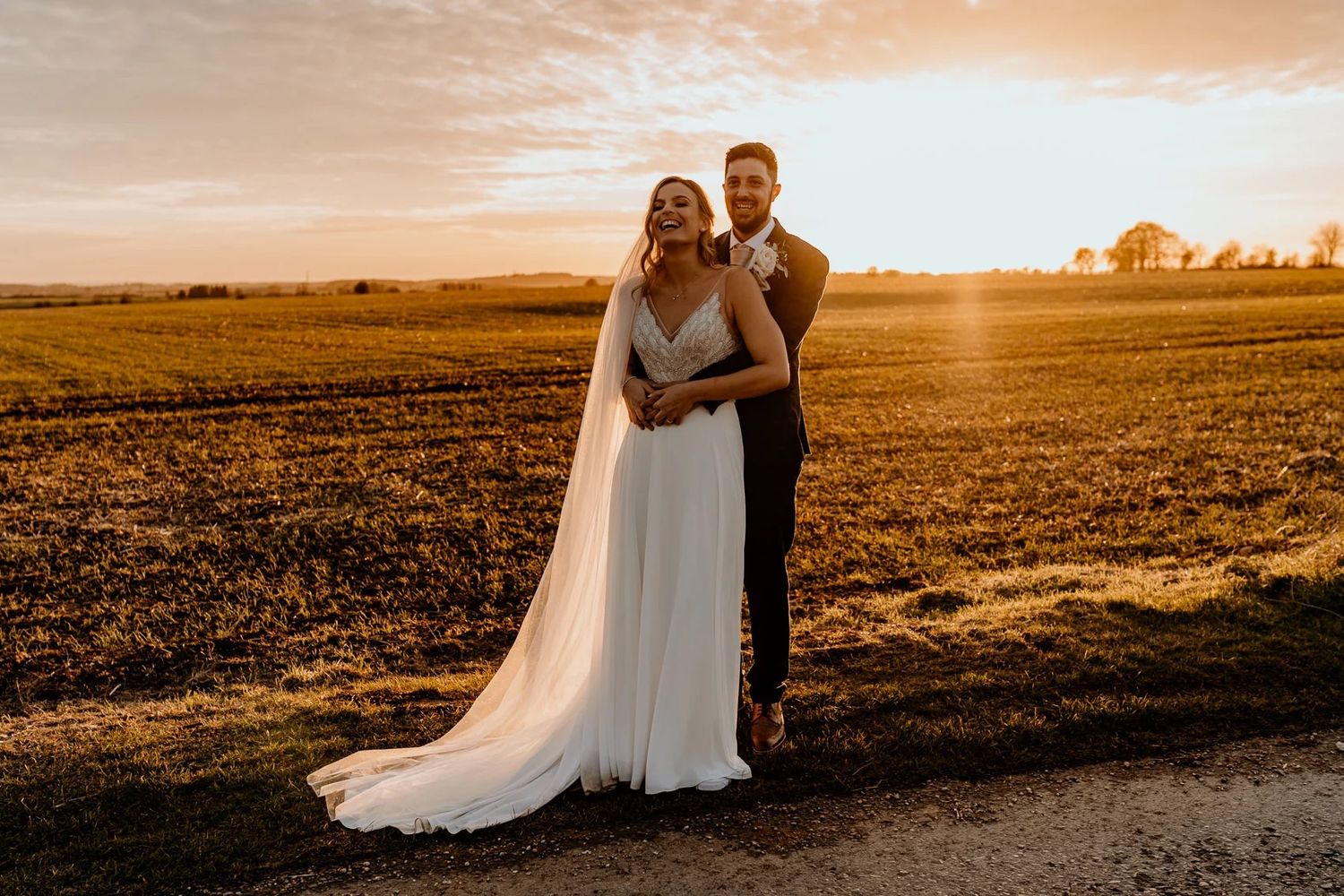 A couple in wedding attire stand together at sunset in a golden field as the sun creates a warm glow on the horizon.