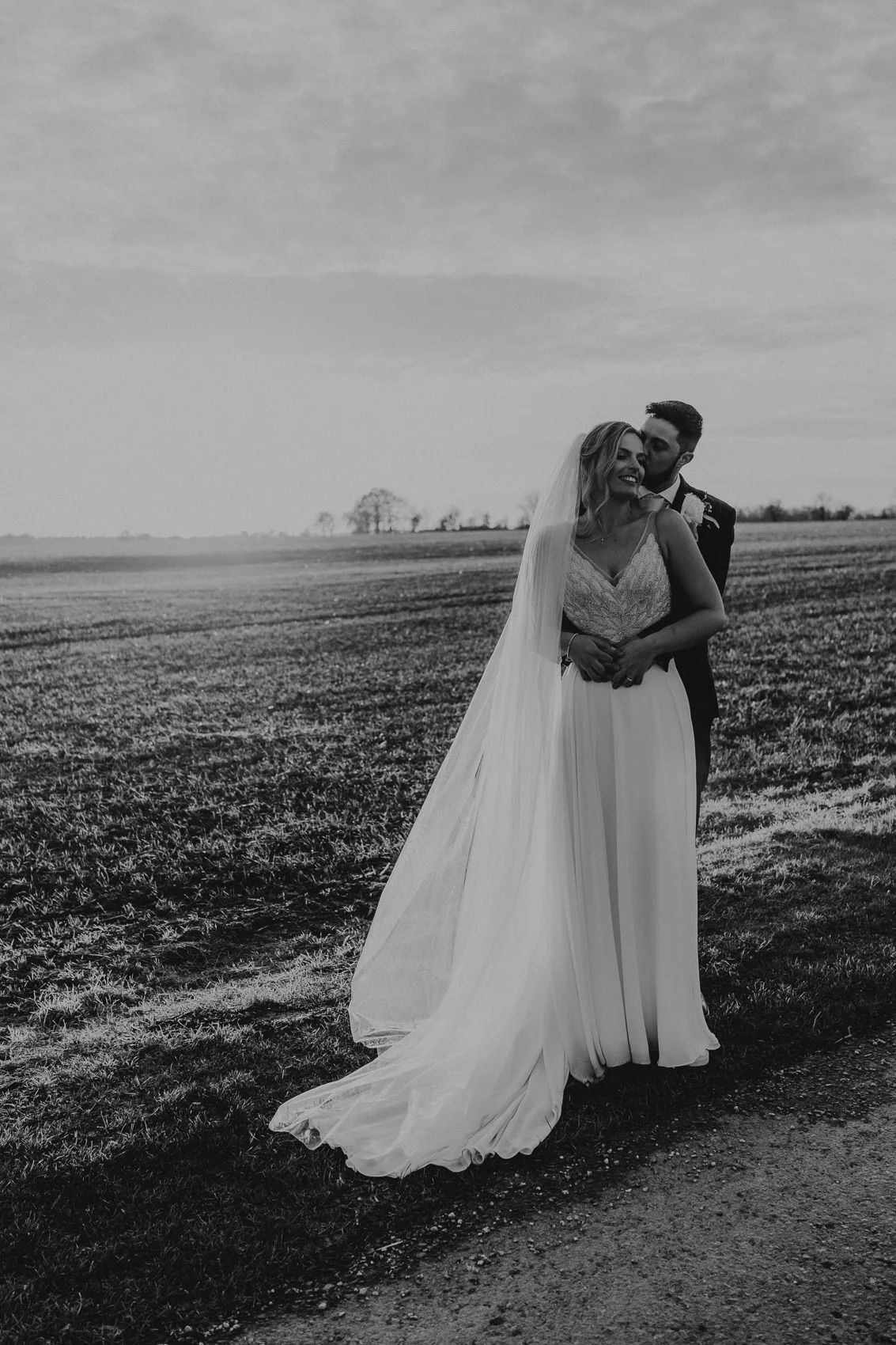 A romantic black and white photo of newlyweds embracing in an open field with flowing wedding dress details.