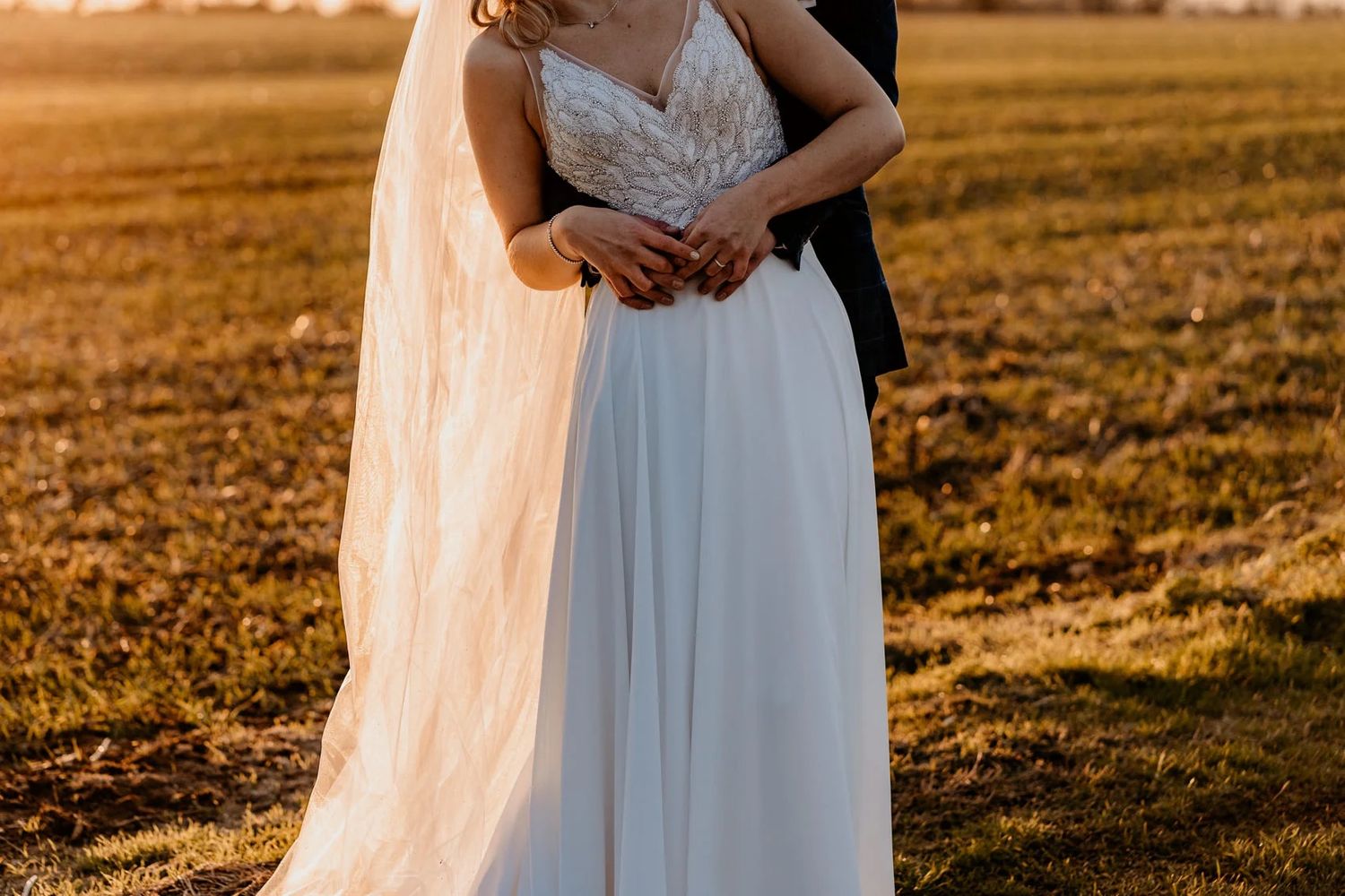 Two elegant figures stand together in a golden field bathed in warm sunset light, flowing white dresses catching the breeze.