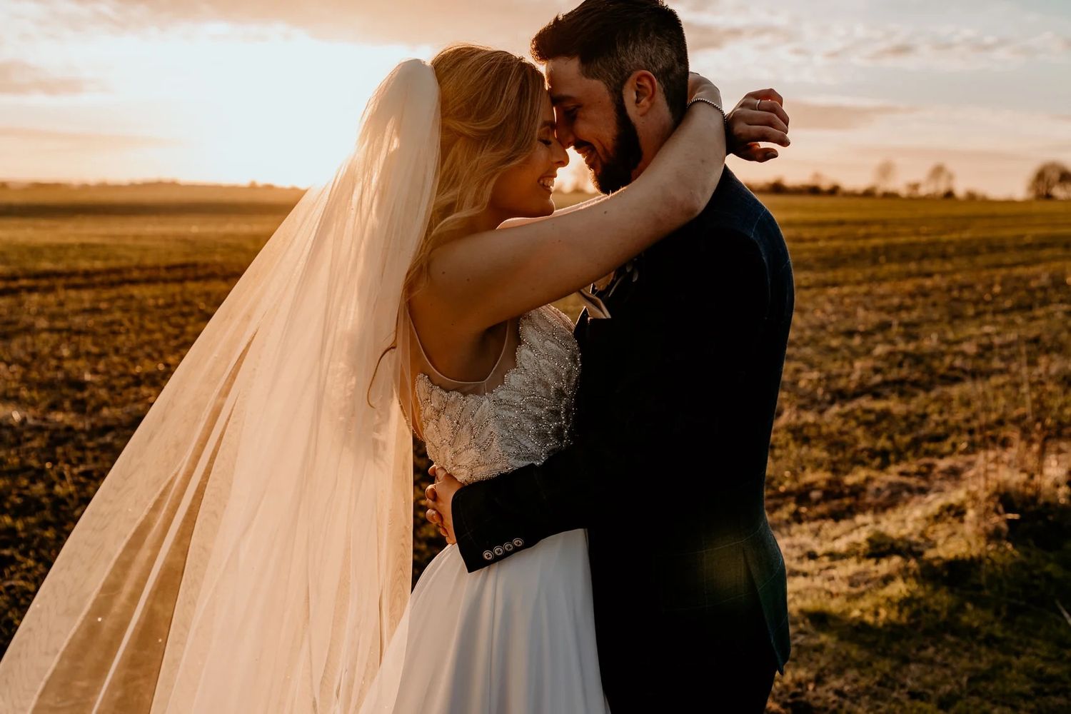 A couple in wedding attire embraces in a golden field at sunset with a long flowing veil.